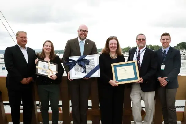 Dignitaries and Museum leadership stand aboard Mayflower II holiding commemorative objects.