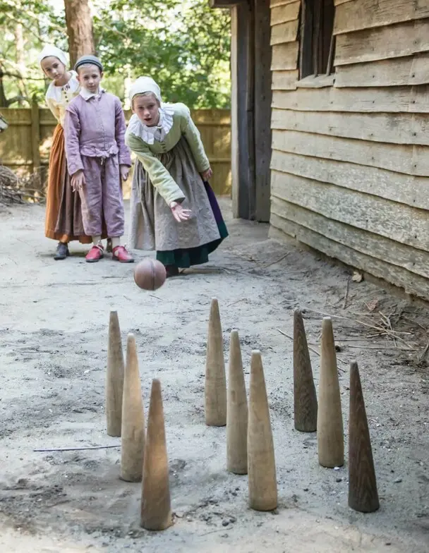 Pilgrim children play a bowling game near a grey, clapboard house.