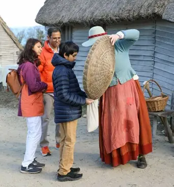 A family of three examines a basket held up by a Pilgrim woman in the 17th-Century English Village.