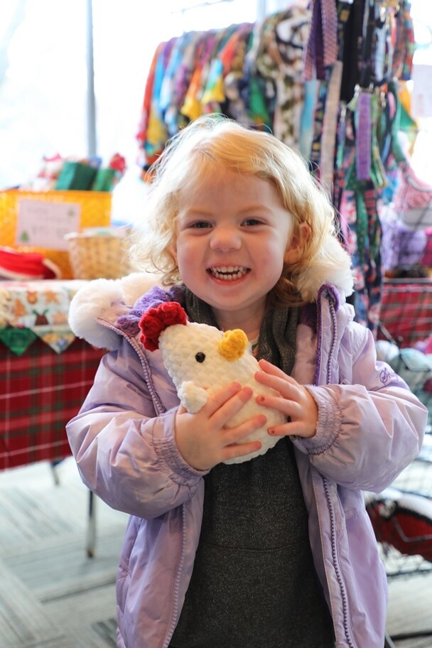 A young, blonde girl smiles as she holds a hand-made stuffed chicken at the Winter Fair.