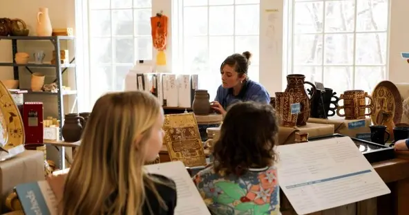 An artisan works to create pottery in the Craft center while two young guests observe her.