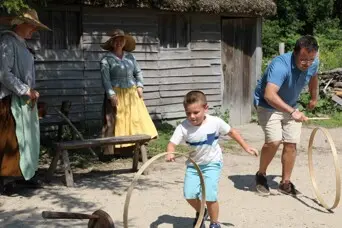 A young boy and his father play a game with wooden hoops while two Pilgrim women watch on.