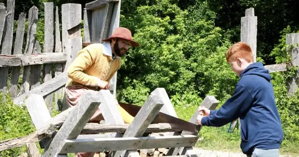 A modern schoolchild helps a Pilgrim saw wood