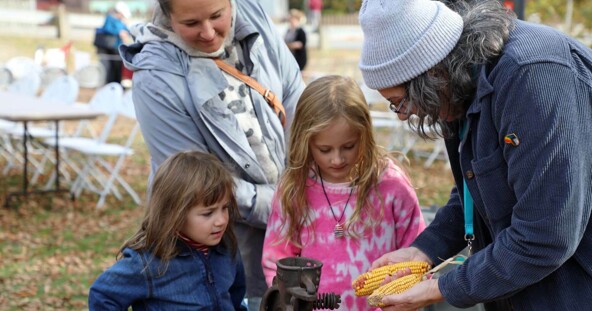Plimoth Grist Mill Educator shows dried corn to two young girls as their mother watches.