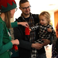 Santa's elf gifts a red velvet bag containing a magical bell to a child being held by their father.