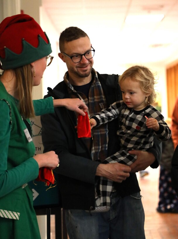 Santa's elf gifts a red velvet bag containing a magical bell to a child being held by their father.