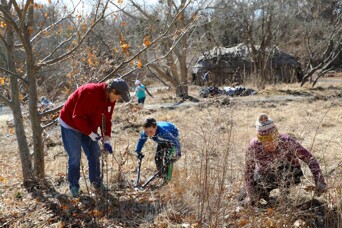 Three volunteers of varying ages work to weed the garden on the Historic Patuxet Homesite. The wetu is behind them in the distance.