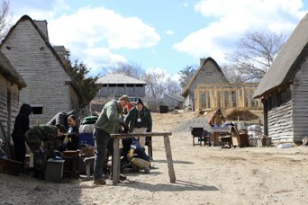 Volunteers congregate around furniture placed in front of a gray, shingled house in the 17th-Century English Village.