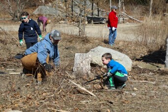 A father weeds the garden on the Historic Patuxet Homesite with his young son. Three volunteers are behind them at varying distances.