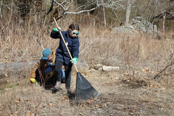 A young girl rakes leaves on the Historic Patuxet Homesite. A staff member crouches to weed behind her.