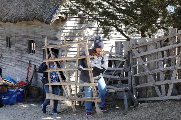 Two volunteers carry a wooden fence panel in the 17th-Century English Village.