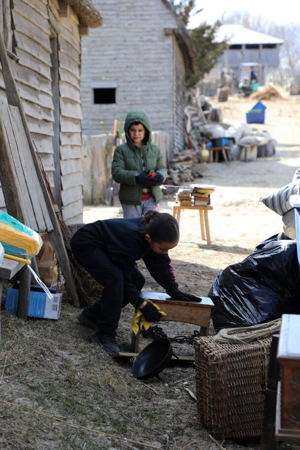 A young girl polishes wooden furniture in front of a Pilgrim house as another young volunteer looks on.