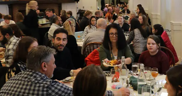 Guests sit at round tables with white tablecloths enjoying Thanksgiving dinner.
