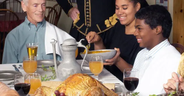 Guests seated at a table enjoy thanksgiving food. A Pilgrim stands behind them.