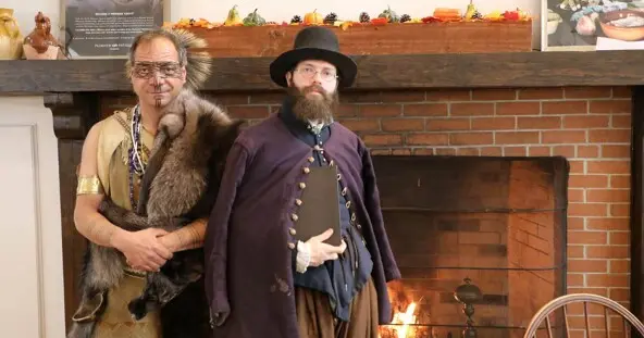 Indigenous Educator dressed in regalia and Educator dressed as a Pilgrim stand in front of a brick fireplace.