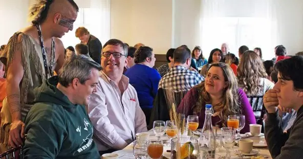 Indigenous Educator dressed in regalia speaks with guests seated for Thanksgiving dinner.