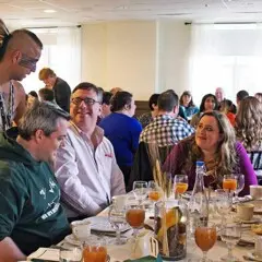 Indigenous Educator dressed in regalia speaks with guests seated for Thanksgiving dinner.