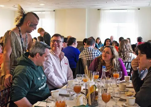 Indigenous Educator dressed in regalia speaks with guests seated for Thanksgiving dinner.