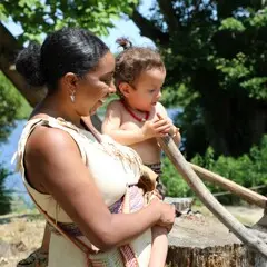 A Museum Educator dressed in Wampanoag regalia holds a toddler boy in regalia has he tries to scrape the mishoon on the Homesite.