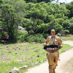 Museum Educator carries a wooden bowl filled with clams through the planting field on the Historic Patuxet Homesite.