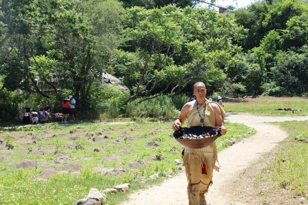 Museum Educator carries a wooden bowl filled with clams through the planting field on the Historic Patuxet Homesite.