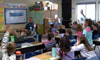 A Museum Educator visits an elementary school classroom. Children raise their hands to ask questions.
