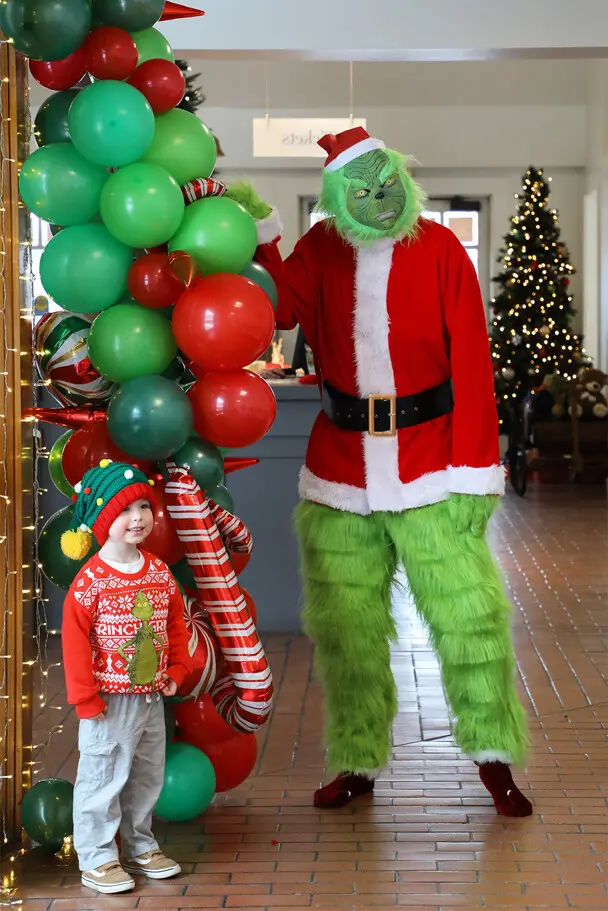 Grinch poses with a young Museum guest in front of a green and red balloon arch.