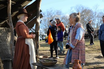 Guest speaks with Pilgrim woman in an outdoor setting during late fall.