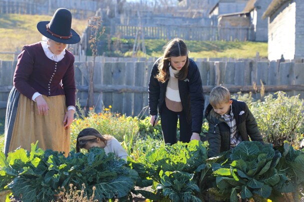 Three children help a Pilgrim woman garden in the English Village.