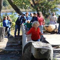 A young, blonde boy in a red jacket runs in a large mishoon. Children behind him enjoy the Historic Patuxet Homesite.