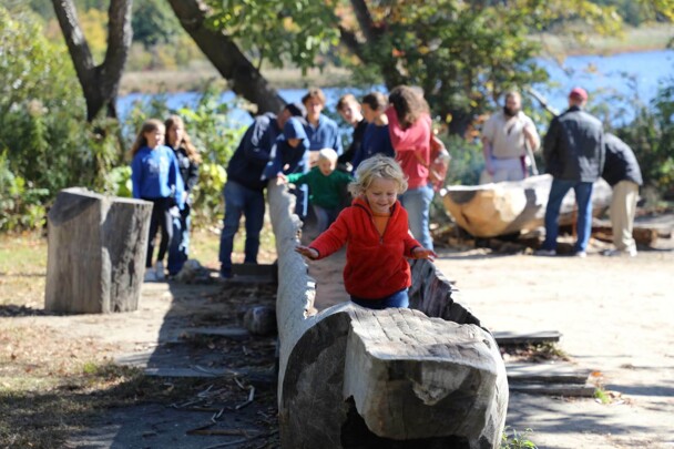 A young, blonde boy in a red jacket runs in a large mishoon. Children behind him enjoy the Historic Patuxet Homesite.