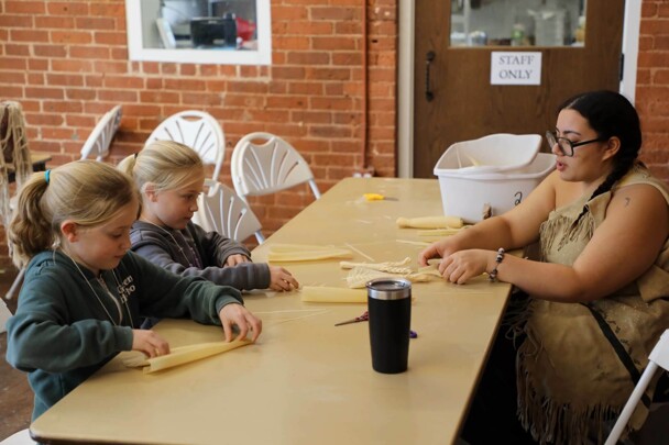 Museum Educator, dressed in Wampanoag regalia, instructs two young, blonde girls, in making cornhusk dolls.