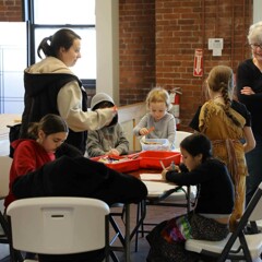 Children seated at a table engage in craft making as two women watch them.