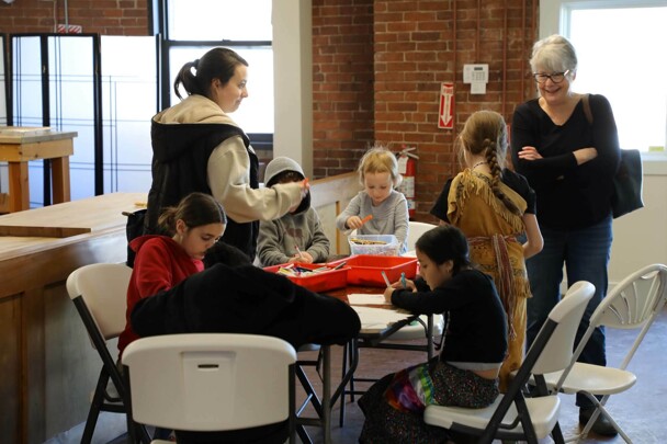 Children seated at a table engage in craft making as two women watch them.