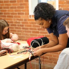 Museum Educator assists a boy with his finger weaving project. A young girl makes her own creation across the table from them.