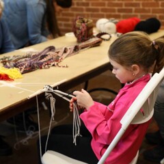 A young girl practices finger weaving at a table with people of all ages who are practicing the craft as well.