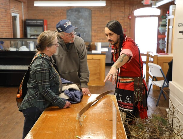 Educator points to an archery bow placed on a wooden table and speaks with an man and woman.