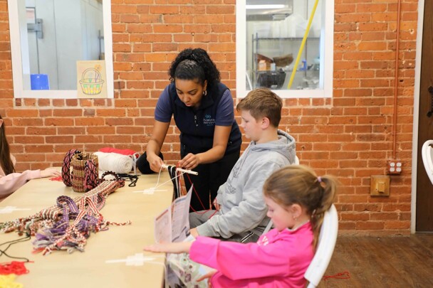 Educator demonstrates finger weaving to a young girl and boy seated at a table in the Craft Center.