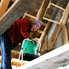 A Museum staffer stands in the eaves of a Pilgrim House with a green bucket as she works to daub walls.