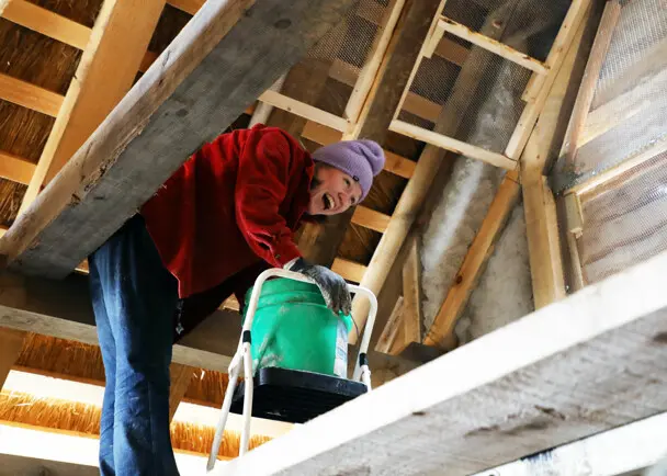 A Museum staffer stands in the eaves of a Pilgrim House with a green bucket as she works to daub walls.