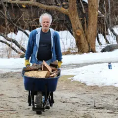 Museum staff moves a wheel barrel filled with wood across the Historic Patuxet Homesite. Snow covers the grounds and the brown winter wetu is in the background.