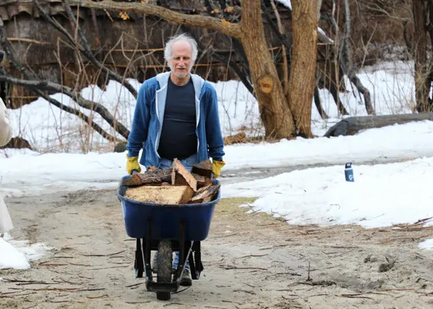 Museum staff moves a wheel barrel filled with wood across the Historic Patuxet Homesite. Snow covers the grounds and the brown winter wetu is in the background.