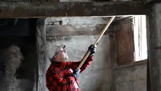 A volunteer dressed in a red flannel shirt raises a wooden broom to sweep above a window inside a Pilgrim house.