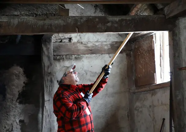 A volunteer dressed in a red flannel shirt raises a wooden broom to sweep above a window inside a Pilgrim house.
