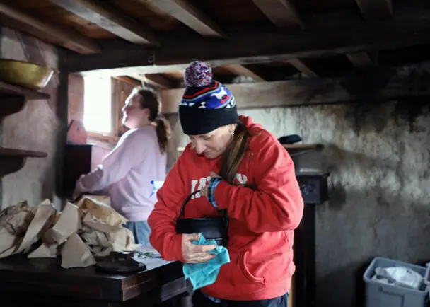 A volunteer in a red sweatshirt and winter hat holds a black pot that she is polishing inside a Pilgrim House.