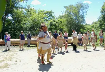 Educators in regalia demonstrate a traditional Wampanoag dance on the Historic Patuxet Homeiste before a large mishoon. Guests gather to watch and learn.