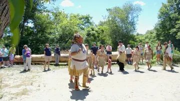 Educators in regalia demonstrate a traditional Wampanoag dance on the Historic Patuxet Homeiste before a large mishoon. Guests gather to watch and learn.