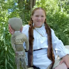 Girl wearing Wampanoag reglia hold up a traditional doll on the Historic Patuxet Homesite