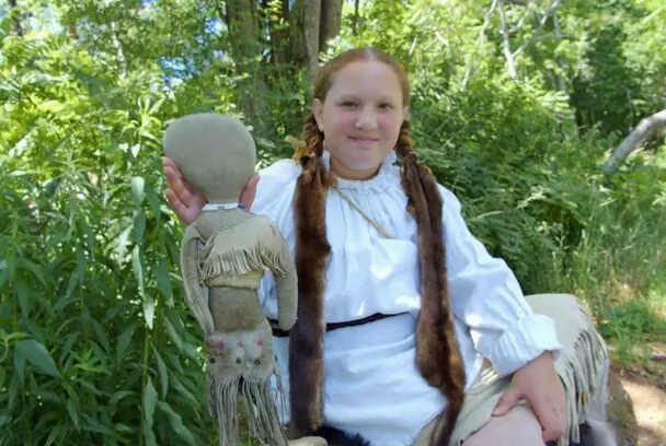 Girl wearing Wampanoag reglia hold up a traditional doll on the Historic Patuxet Homesite