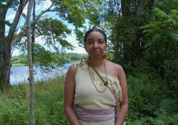 Museum Educator dressed in Wampanoag regalia surrounded by greenery